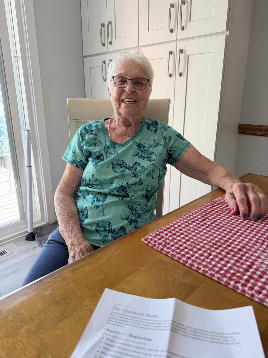 A grandmother at her kitchen table, mid-interview, with the Tend & Tell question bank in front of her